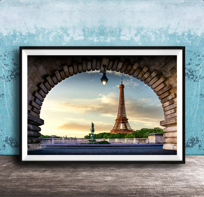 A framed photograph of the Eiffel Tower in Paris, France, with the tower visible through an archway. The sky is blue with clouds, and the sun is setting, creating a picturesque scene.