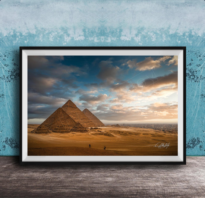 two large sandstone pyramids against a cloudy sky, with a few people walking in the foreground.