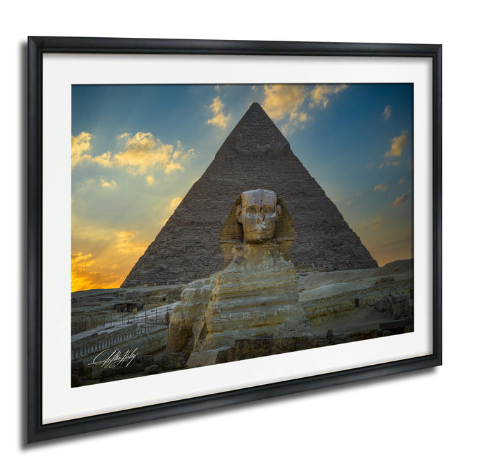 a large stone statue of a man with a headdress, standing in front of the iconic Great Pyramid of Giza in Egypt. The sky above the pyramid is filled with clouds, creating a dramatic backdrop for the scene. The photograph is framed and appears to be a high-quality image of the Great Pyramid and the Sphinx.