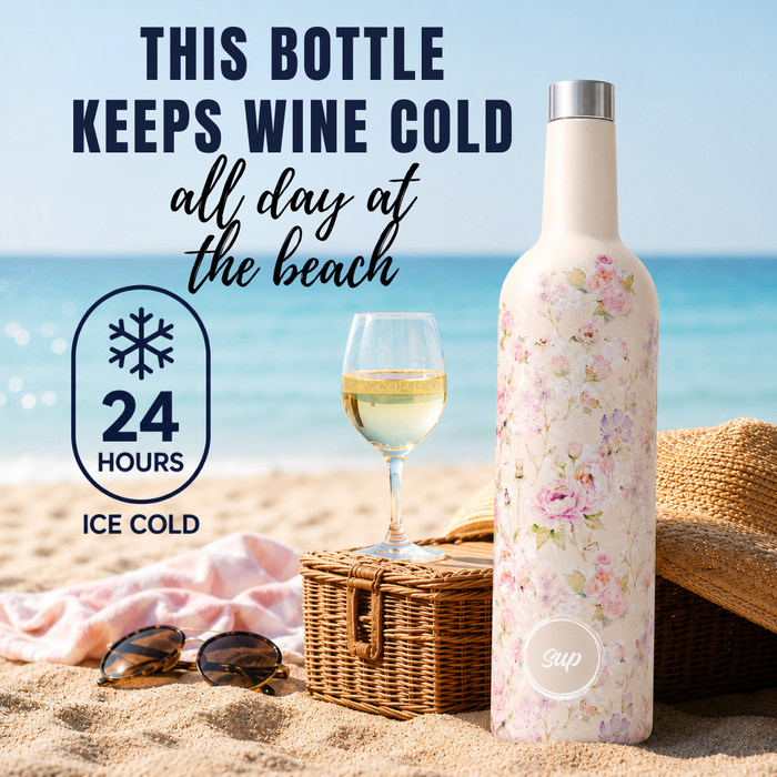 A floral insulated wine bottle stands on a sandy beach next to a glass of white wine, a wicker picnic basket, sunglasses, and a straw hat, with text reading 'THIS BOTTLE KEEPS WINE COLD all day at the beach' and '24 HOURS ICE COLD'.
