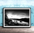 A black and white landscape photograph depicting a serene lake surrounded by mountains and pine trees, with a cloudy sky overhead.