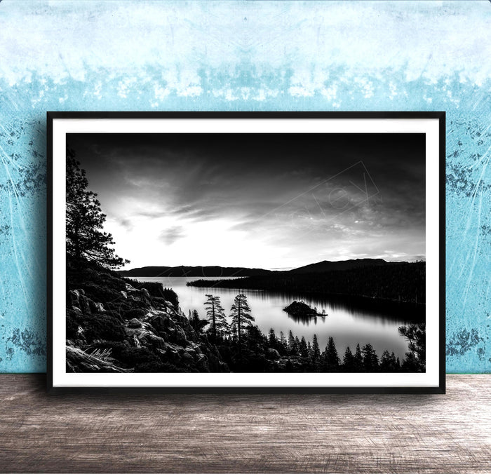 A black and white landscape photograph depicting a serene lake surrounded by mountains and pine trees, with a cloudy sky overhead.