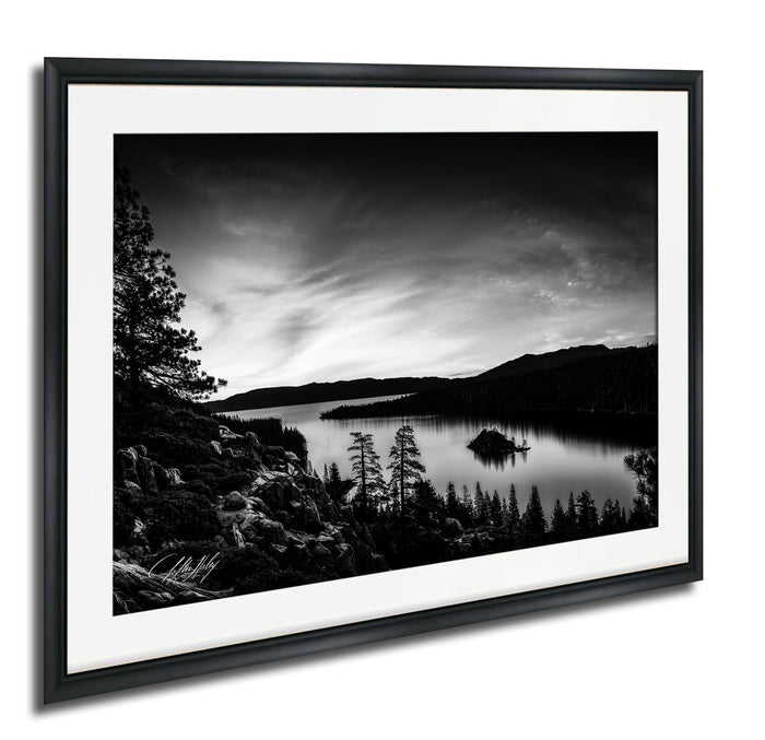 A black and white landscape photograph depicting a serene lake surrounded by mountains and trees, with a small island in the center.