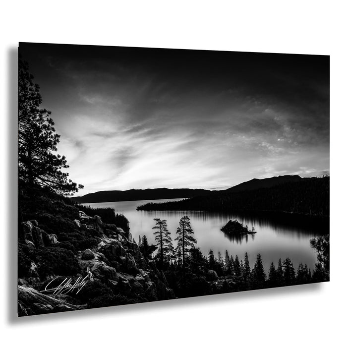 A black and white landscape photograph depicting a serene lake surrounded by mountains and pine trees, with a small island in the center.