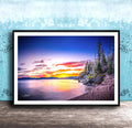 A framed photograph depicting a serene landscape with a calm lake reflecting the vibrant colors of a sunset sky, surrounded by trees and mountains.