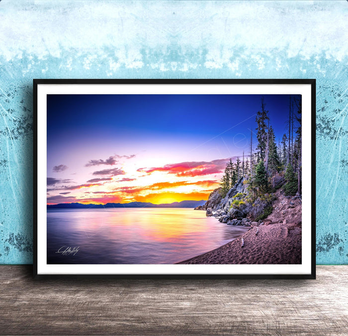 A framed photograph depicting a serene landscape with a calm lake reflecting the vibrant colors of a sunset sky, surrounded by trees and mountains.
