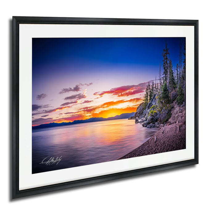 A framed photograph depicting a serene landscape with a calm lake reflecting the vibrant colors of a sunset sky, surrounded by trees and mountains.