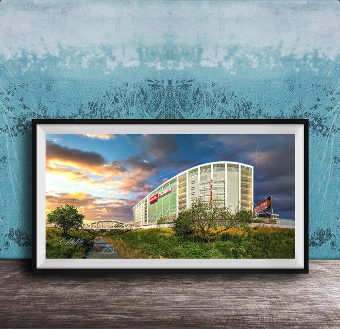 A framed photograph of a large building with a curved roof, surrounded by trees and a river, with a colorful sunset sky in the background.