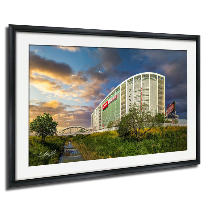 A framed photograph depicting a large building with a prominent red sign, set against a backdrop of a colorful sunset sky and lush greenery.