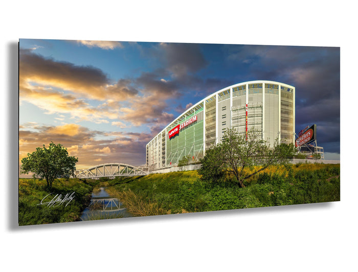A large stadium with a curved roof and a bridge in the foreground, set against a colorful sky with clouds.