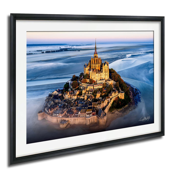 a picturesque island with a castle on top, surrounded by a body of water. The castle is illuminated, creating a warm and inviting atmosphere. The island is nestled in a bay, with the water surrounding it. The sky above is a beautiful shade of blue, with a few clouds scattered across it. The image is framed, adding a touch of elegance to the scene.