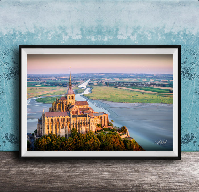 a large, ornate cathedral with a tall spire, situated on a cliff overlooking a body of water. The cathedral is surrounded by lush greenery, creating a picturesque scene. The photograph is framed and displayed on a wooden surface, with a blue wall in the background.