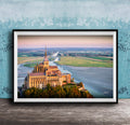 a large, ornate cathedral with a tall spire, situated on a cliff overlooking a body of water. The cathedral is surrounded by lush greenery, creating a picturesque scene. The photograph is framed and displayed on a wooden surface, with a blue wall in the background.