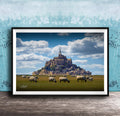 A framed photograph depicting a pastoral scene with a large castle or hill in the background, surrounded by a grassy field where a herd of sheep are grazing.