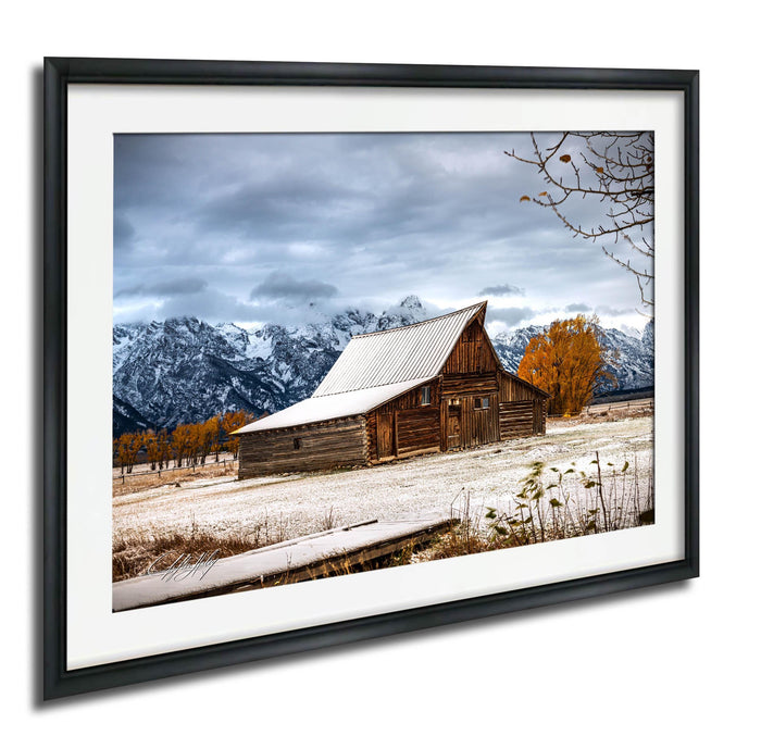 A rustic wooden barn with a sloping roof stands in a snowy field, surrounded by snow-capped mountains and a cloudy sky.