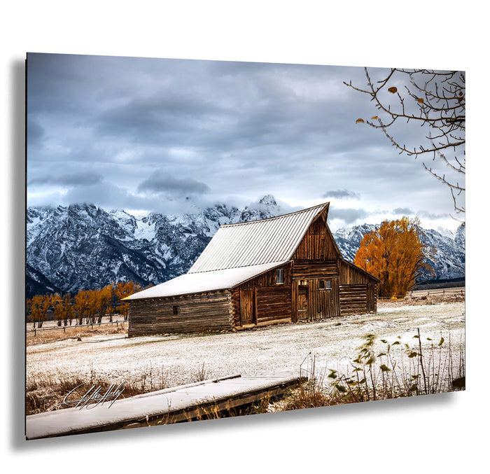 A rustic wooden barn stands in a snowy field, with snow-capped mountains in the background and a few trees with orange foliage.