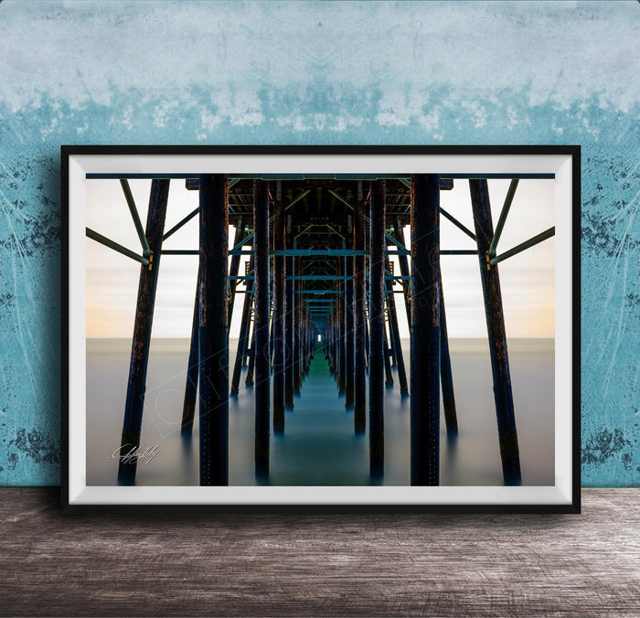 The image is a framed photograph of a long pier with multiple wooden columns extending into the distance, creating a sense of depth and perspective.