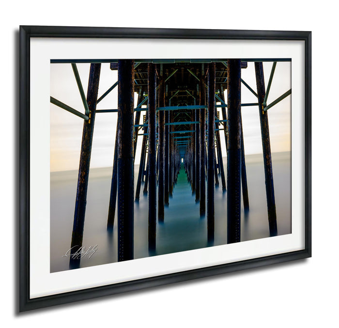 The image is a framed photograph of a long pier with wooden columns extending into the distance, creating a sense of depth and perspective.