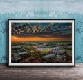 The image is a framed aerial view of a cityscape at sunset, with the sky filled with clouds and the sun setting over the horizon.