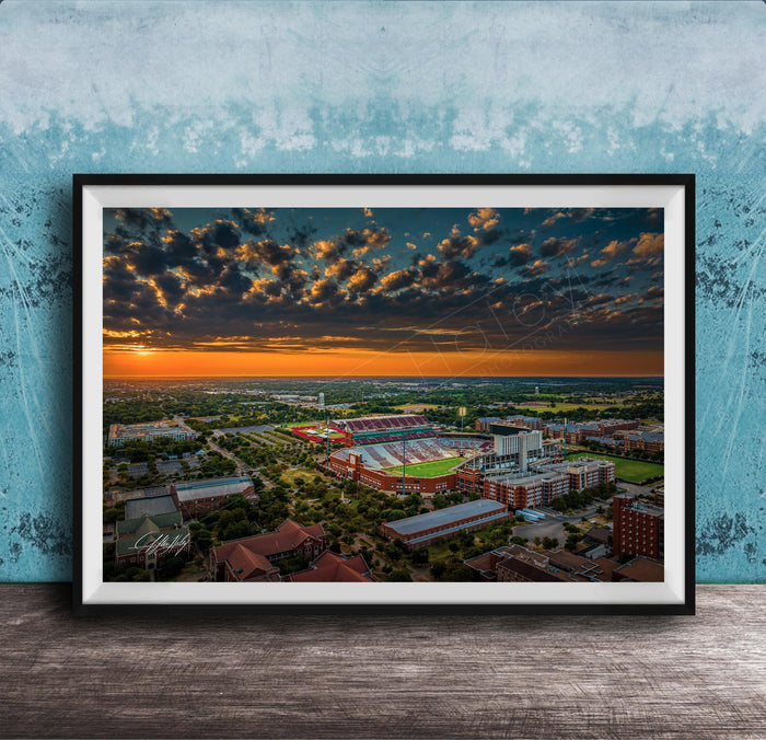 The image is a framed aerial view of a cityscape at sunset, with the sky filled with clouds and the sun setting over the horizon.