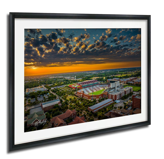 The image is an aerial view of a large sports stadium surrounded by a cityscape, with a dramatic sky in the background featuring clouds and a colorful sunset.