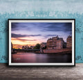 a picturesque cityscape at sunset, with a river running through the center and a row of buildings lining the riverbank. The sky is filled with warm, colorful hues, creating a serene and tranquil atmosphere. The buildings, with their unique architectural styles, add to the charm of the scene. The image is framed and displayed on a wooden surface, enhancing the overall aesthetic of the setting.