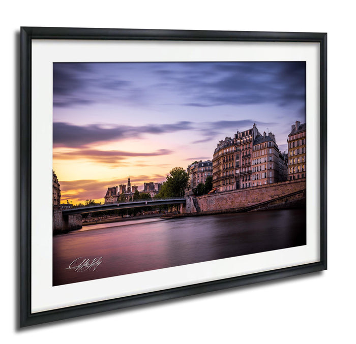 a picturesque cityscape with a river running through it, featuring a bridge and buildings along the riverbanks. The sky is painted in warm hues of orange and pink, creating a serene and tranquil atmosphere. The image is framed and appears to be a high-quality, professional-looking photograph.