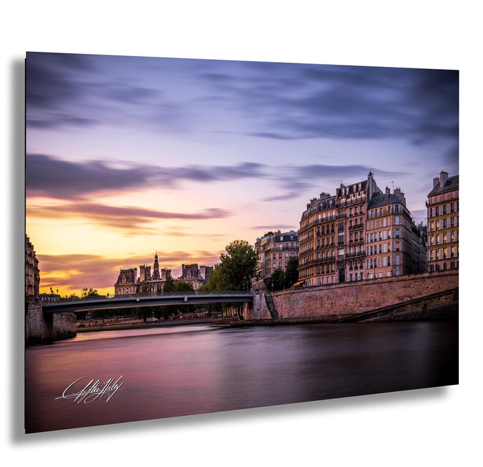 a picturesque cityscape with a river running through it, featuring a bridge and buildings with ornate architecture. The sky is filled with clouds, creating a dramatic backdrop for the scene. The colors in the image are vibrant, with the warm hues of the setting sun casting a golden glow over the city.