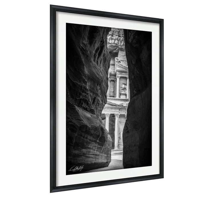 A black and white photograph of a narrow passage between two large rock formations, with the entrance to a building visible through the opening.