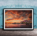 A framed photograph depicting a serene sunset over a wooden pier, with a small building on the pier and a calm body of water in the foreground.