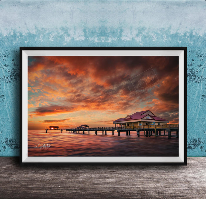A framed photograph depicting a serene sunset over a wooden pier, with a small building on the pier and a calm body of water in the foreground.