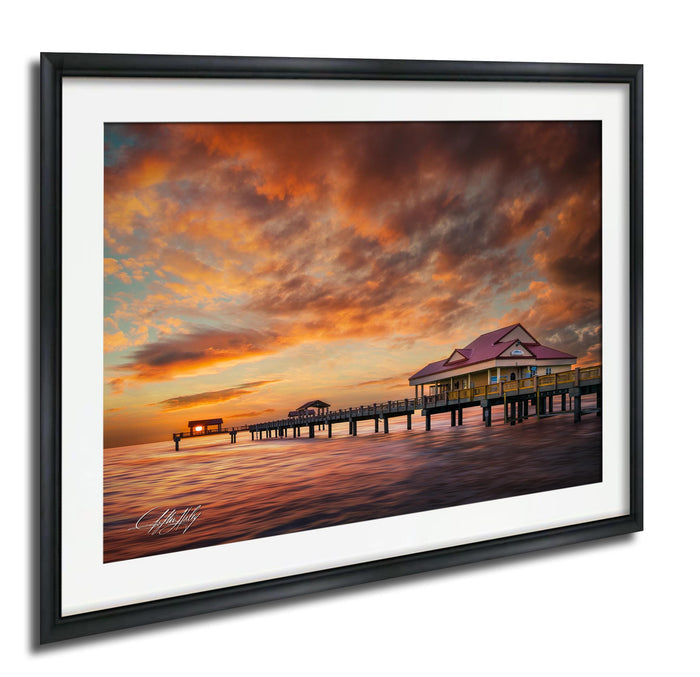 A framed photograph depicting a serene sunset over a wooden pier with a small building on it, set against a vibrant sky filled with clouds.