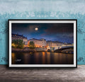 a picturesque night scene of a city with a bridge and buildings illuminated by lights, with a full moon visible in the sky.