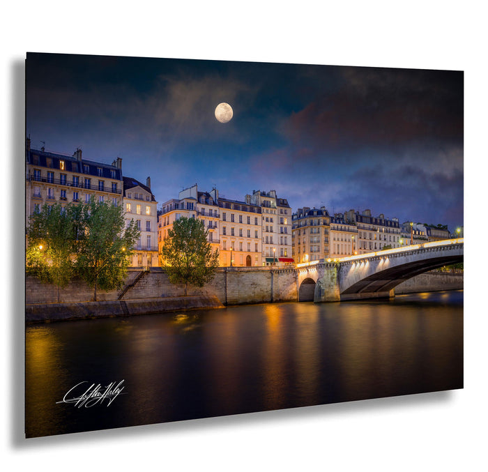a serene night scene in Paris, France, with the Seine River flowing through the cityscape. The moon is visible in the sky, casting a soft glow over the illuminated buildings and bridge. The photograph is taken from a distance, providing a panoramic view of the city's architectural beauty and the tranquil waters of the river.