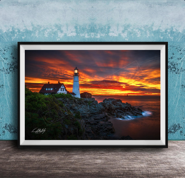 A framed photograph depicting a lighthouse on a rocky cliff at sunset, with the sky filled with vibrant orange and red hues.