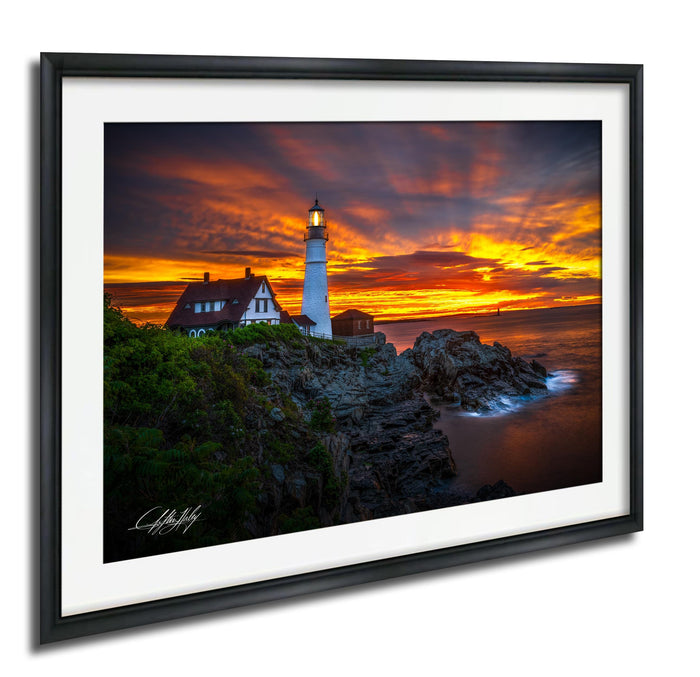A framed photograph depicting a lighthouse on a rocky cliff at sunset, with a vibrant sky and ocean in the background.