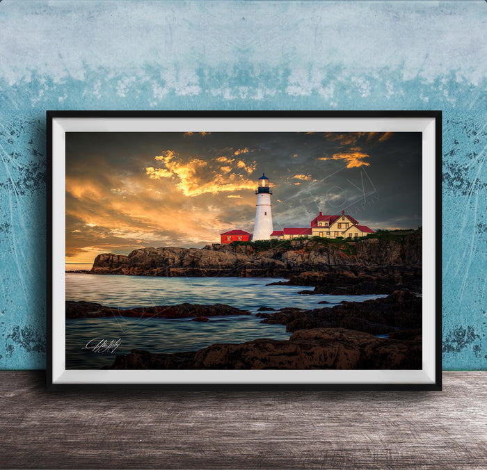 A framed photograph depicting a lighthouse on a rocky coastline at sunset, with the sky filled with clouds and the ocean in the background.