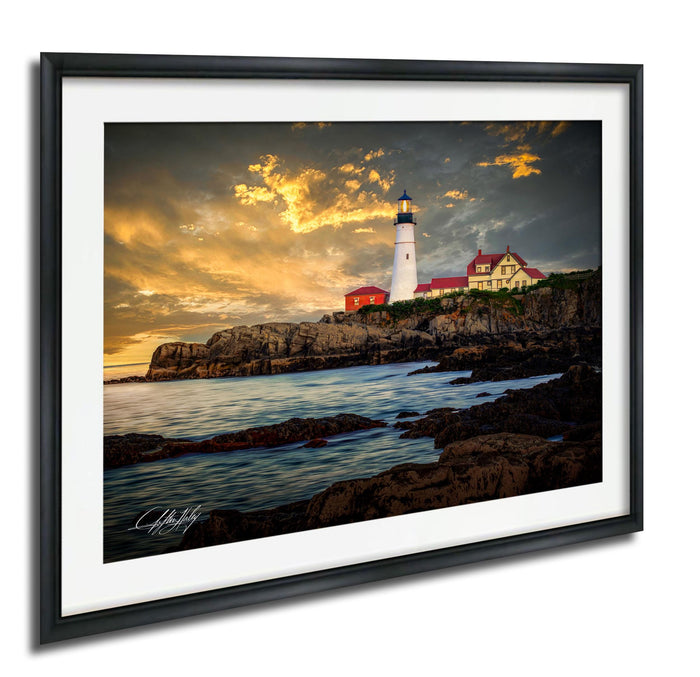 A framed photograph depicting a lighthouse on a rocky coastline at sunset, with the sky filled with clouds and the ocean in the background.