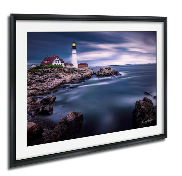 A framed photograph depicting a lighthouse on a rocky shore, with the ocean and a cloudy sky in the background.