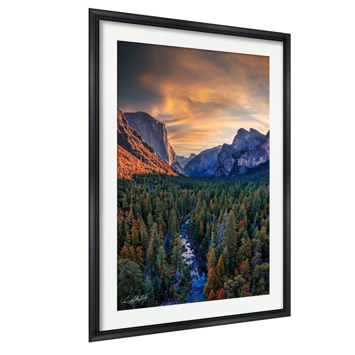 The image is a framed photograph of a scenic mountain landscape with a river flowing through a forested valley. The mountains are covered in snow, and the sky is filled with vibrant colors, creating a breathtaking natural scene.
